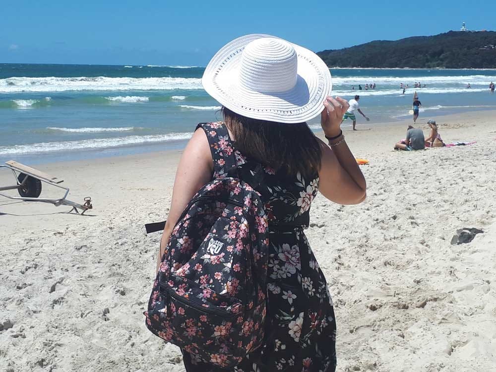 chica morena de espaldas con un sombrero blanco y la mochila de AUssieYouTOO. Está en la playa